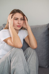 Young woman holding her head, frustrated, sitting on sofa in modern living room during daytime