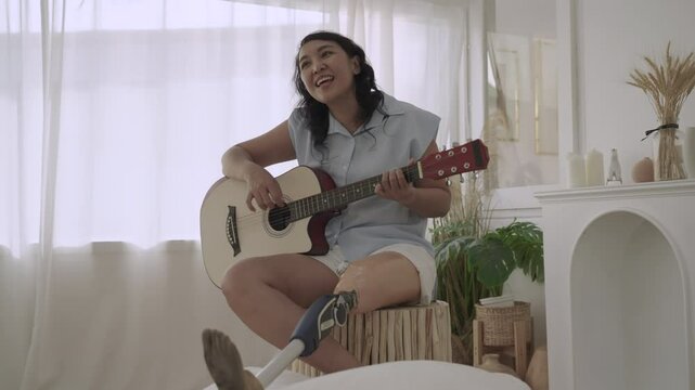 Happy Asian woman with prosthetic leg disability sitting playing acoustic guitar and singing having fun and cheerful in the living room at home. Positive life of a female her leg prosthesis equipment.