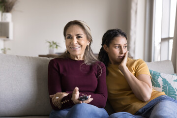 Happy senior Latin mom and bored adult daughter disagree on channel choice watching movie, show, series on TV together, sitting close on home sofa, holding remote control