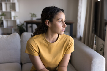 Serious thoughtful attractive 20s Latin woman looking at window away in deep thoughts, sitting on sofa, thinking on problem solving at home. Concerned pensive Hispanic young adult girl casual portrait