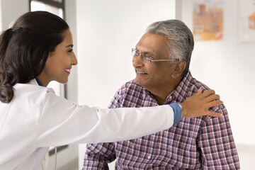 Positive female doctor or nurse giving psychological support to smiling Indian senior male patient...