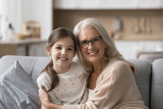 Loving grandmother cuddling her little 6s adorable granddaughter, smiling, posing together for camera sit on couch at home. Multigenerational relatives portrait, family ties, understanding and bonding