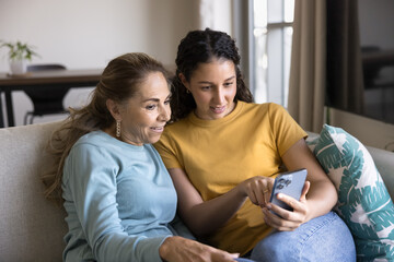 Young adult Latin daughter teaching senior mother to use smart home application on mobile phone,...