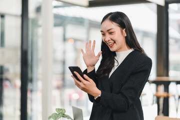 Fototapeta premium Smiling woman in black blazer waving at smartphone, enjoying video call in modern office space, showcasing happiness and connection
