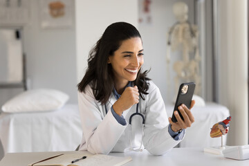 Smiling Latina doctor in white coat holds smartphone look at screen device lead online videocall...