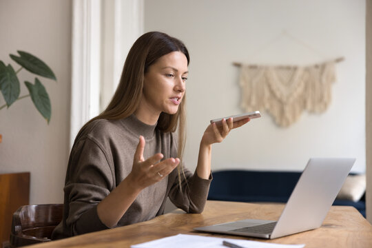 Young businesswoman sitting at table with laptop, using smartphone, recording voicemail, audio message or interacting with voice assistant, engaged in remote communication. Innovative voice technology