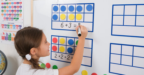 Little girl standing in classroom near white magnetic board, smiling, solving math problems using frames and counters