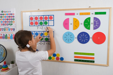 Little girl standing in classroom near white magnetic board, smiling, solving math problems using...