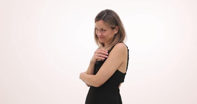 Shy woman embraces own arms during conversation on white background. Smiling female in black cocktail dress experiences satisfaction with compliment