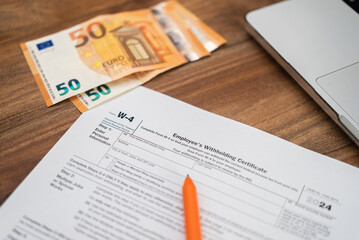 Obraz premium Employee filling out IRS form W-4 with euros and a laptop on a wooden desk. Payroll, tax withholding, and personal finance management concept 