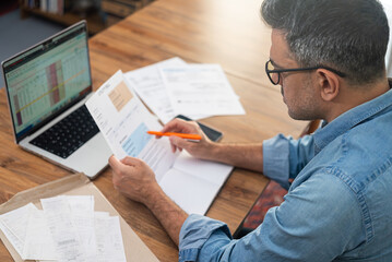 Man reviewing utility bills at home with laptop and documents on wooden table. Concept of household expenses, family budget, and financial planning 