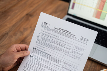 Close-up of a hand holding a W-9 tax form with a laptop in the background. Tax preparation and...