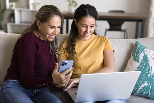 Positive elder Latin mom and happy young daughter woman using gadgets simultaneously at home, watching content on laptop and smartphone, sitting on couch with digital devices, laughing