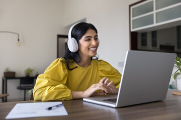 Positive Indian student girl in big wireless headphones watching learning webinar at laptop,...