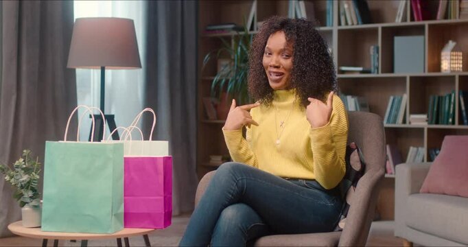 Close up on a young african american blogger making fashion video with packages for subscribers. Cute woman records clothes video smiling widely, talking, moving hands and showing white shirt.
