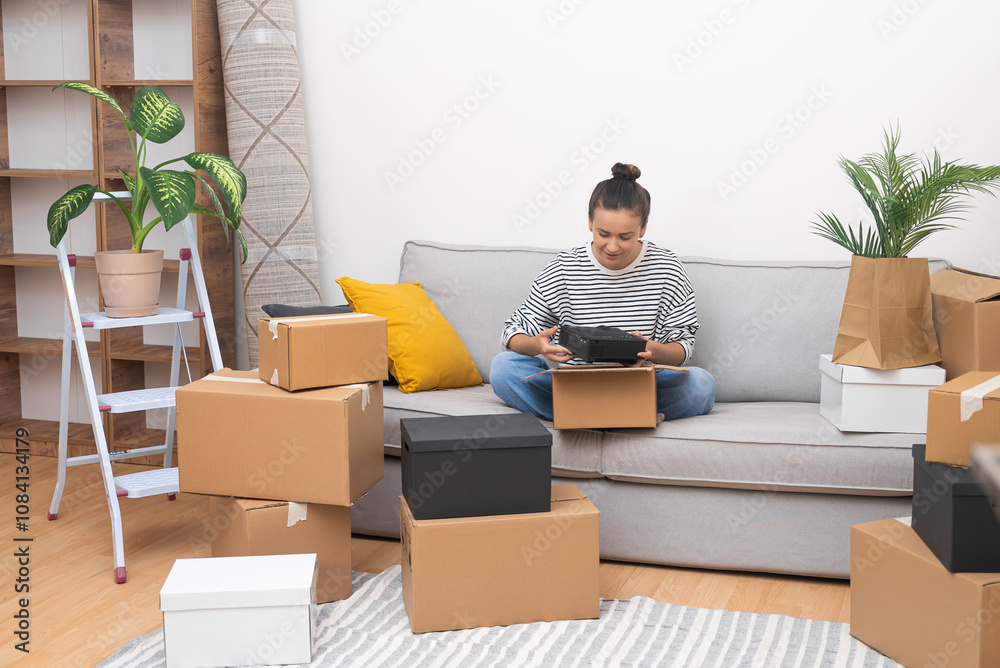 Poster smilling lady with bun o head with delighted expression sits on sofa collecting cardboard boxes for 