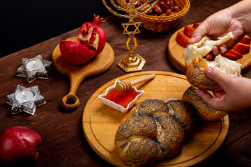 Female hands breaking challah on the table meal in Rosh Hashanah next to apple honey menorah...