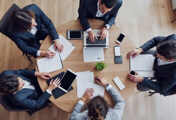 Top view of a diverse group of business professionals in a meeting, discussing strategy, and using technology around a round office table.