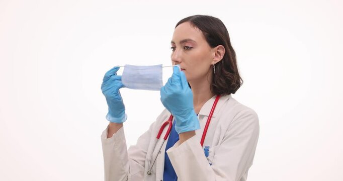 Young woman doctor in lab coat puts on medical mask. Female physician with stethoscope prepares for patient examination on white background