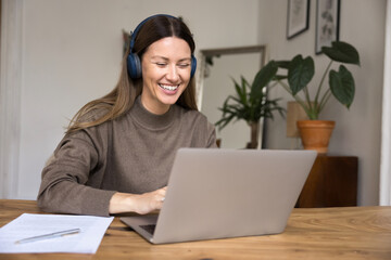Smiling pretty woman sit at desk, wear headphones listen audio course, e-learning, studying, gain...
