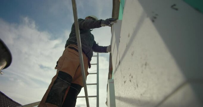 Placing insulation panel on exterior wall, construction worker on scaffold using hands for precise positioning, safety gear visible, residential building facade