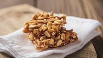 Peanut or Almond Brittle: A piece of golden-brown brittle, sweet and crunchy, placed on a white cloth with a rustic wooden background, evoking a homely, cozy vibe.