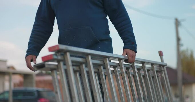 Worker carrying aluminum ladder at construction site, preparing for outdoor task, emphasizing utility and equipment for renovation and construction work