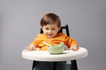 Cute little kid eating healthy baby food from bowl in high chair on light grey background