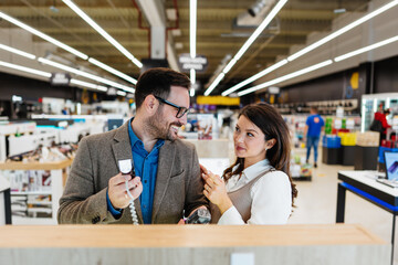Beautiful and happy middle age couple buying consumer tech products in modern home appliances store. They are choosing beard and hair clipper and hairdryer. People and consumerism concept.