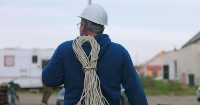 Worker walking with cables on shoulder near outdoor workspace, preparing for electrical work and organizing tools for efficient construction project execution