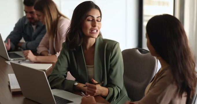 Positive confident beautiful Latin businesswoman talking to female office colleague at workplace in co-working office, explaining task to new employee, intern, speaking to coworker