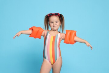Cute little girl in swimsuit with swim armbands on light blue background