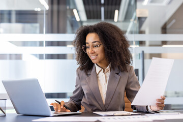 A smiling young African-American woman is sitting at the desk in the office, holding documents and looking at the laptop screen