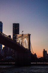 Obraz premium Elegant view of the Brooklyn Bridge illuminated at dusk, set against a soft gradient sky as the city skyline emerges in the background