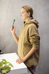 A young woman in a cozy beige hoodie thoughtfully examines her skin in a circular illuminated mirror, standing in a modern minimalist bathroom.