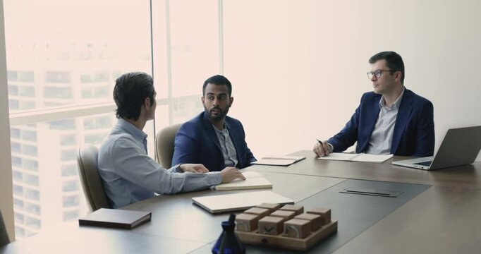 Confident young Indian businessman speaking to listening colleagues on stakeholders meeting, explaining idea for teamwork. Small team of company leaders talking at table