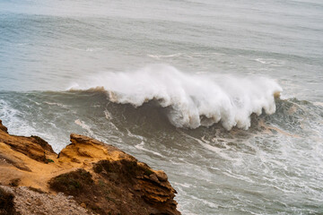 Big wave with white foam hitting the rocky shore at Nazare in Portugal