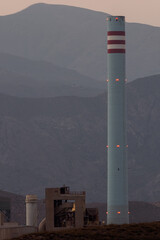 Tall industrial chimney with red and white stripes in front of a mountain landscape during twilight