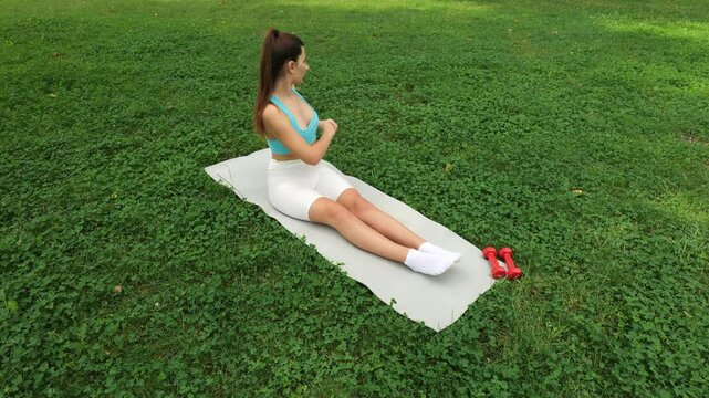 Young athletic woman doing stretching sitting on grey gym mat. Workout on green grass in city park