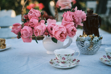 Elegant outdoor tea party scene with pink roses in a white vase, teacups, and chocolates.  Perfect for blogs, magazines, or websites related to tea, spring, or garden parties.