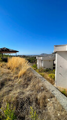 Abandoned structures with overgrown vegetation on a hillside overlooking a scenic coastal city under a clear blue sky, concept of urban decay, redevelopment, and environmental renewal projects.