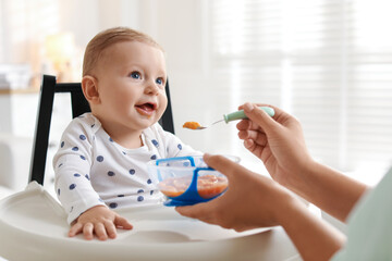 Mother feeding her cute little baby in high chair at home, closeup