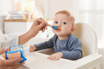 Mother feeding her cute little baby in high chair at home, closeup