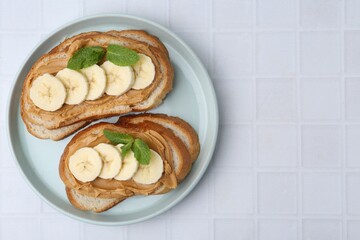 Tasty sandwiches with peanut butter, banana and mint on white tiled table, top view. Space for text