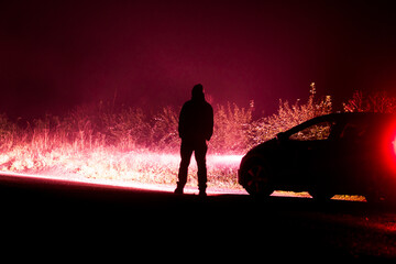 A mysterious hooded figure, silhouetted by car headlights on a country road. With a dark red edit.