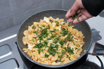 A closeup of a women hand cooking the pasta macaroni or penne pasta in wok pan, pouring spring onions and celery to the pasta macaroni or penne pasta in pan,  cooking pasta homemade.