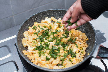 A closeup of a women hand cooking the pasta macaroni or penne pasta in wok pan, pouring spring onions and celery to the pasta macaroni or penne pasta in pan,  cooking pasta homemade.