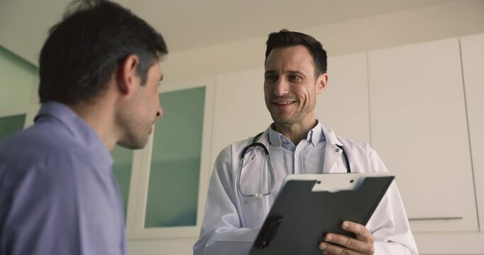 Positive mature Latin doctor holds journal card filling medical form during talk to patient during visit in hospital, pass annual health check-up, share complaint or symptoms, receive professional aid