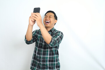 carefree asian man wearing casual plaid shirt laughing while watching video on smartphone isolated on white background