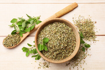 Dried oregano in bowl, spoon and green leaves on wooden table, top view